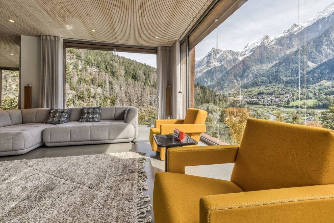 Una sala de estar moderna con un amplio sofá modular, un sillón amarillo y vistas a un paisaje montañoso a través de ventanales de pared a techo.