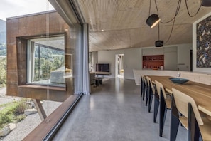 A modern interior with a dining area, kitchen, and large windows offering a view of the outdoors. - Skyfall - Modern Chalet | Sleeps 10 | Unique Design | View (Les Houches)