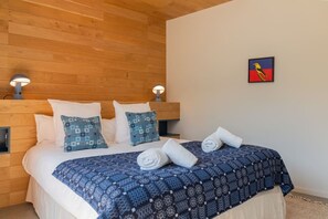 A hotel room with a wooden headboard, a bed with white and blue bedding, two wall-mounted lamps, and a painting on the wall. - Skyfall - Modern Chalet | Sleeps 10 | Unique Design | View (Les Houches)