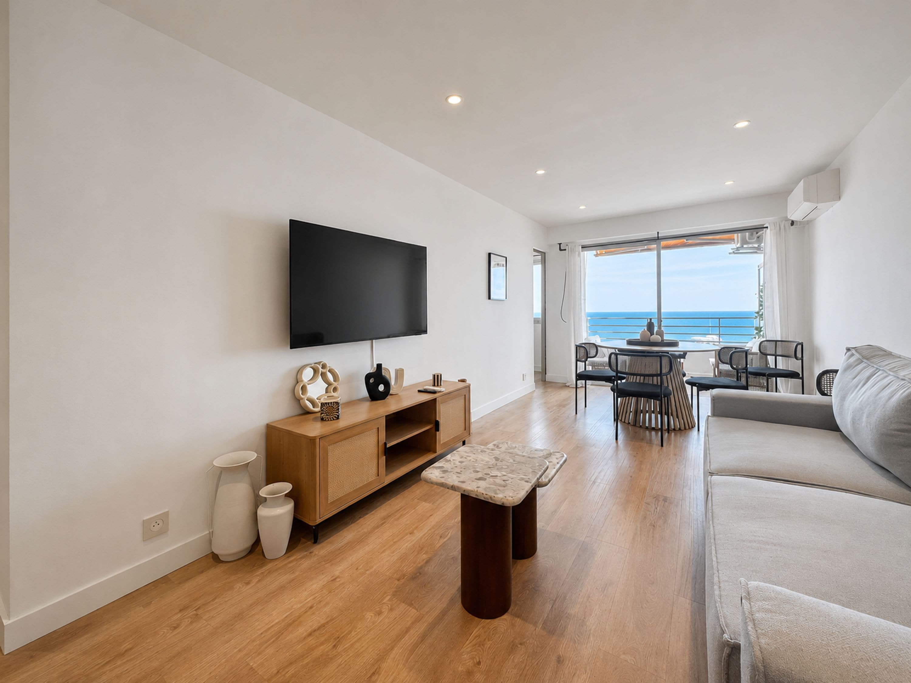 A modern living room with a flat-screen TV, wooden furniture, a sofa, and a dining area with a view of the sea.