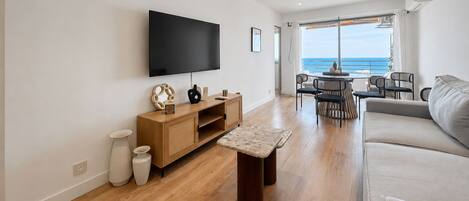 A modern living room with a flat-screen TV, wooden furniture, a sofa, and a dining area with a view of the sea.