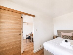 A bedroom with a bed, a bedside table, a nightstand, a wooden door, and a dining area with a table and chairs. - 2BR Sea View Central 1 Min From Monaco (Beausoleil)