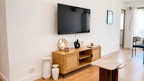 A modern living room with a flat-screen TV mounted on the wall, a wooden cabinet with decorative items, and a small table with a cushion. - 2BR Sea View Central 1 Min From Monaco (Beausoleil)