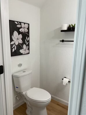 A bathroom with a toilet, a white toilet paper holder, and a black shelf with a roll of toilet paper and a small plant. - Rezidence Townhome at Dignowity (San Antonio)