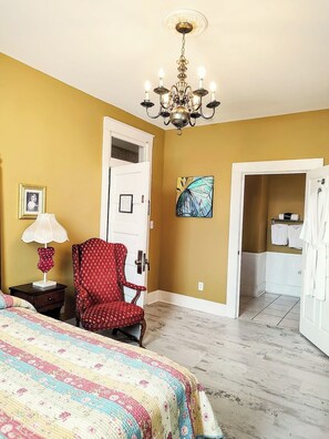 Une pièce comprenant un lit, une chaise, un lustre, une porte et une salle de bain visible à travers une porte ouverte.