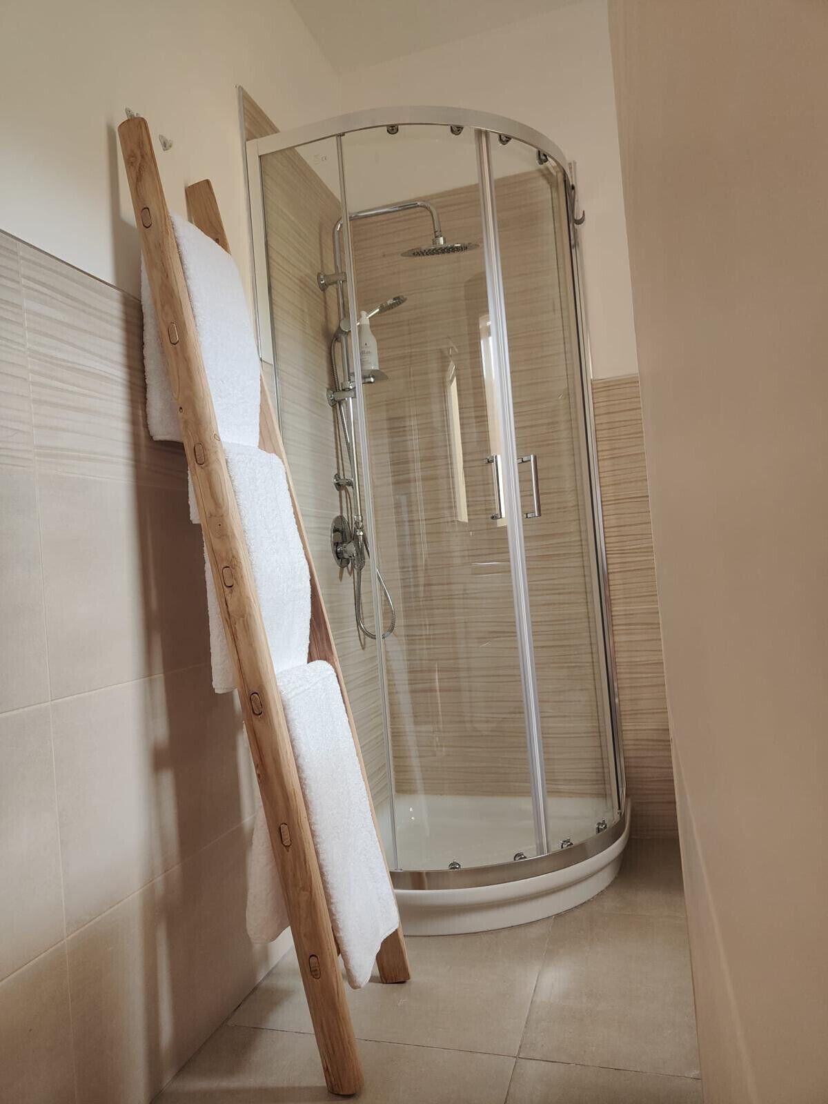 A corner bathroom with a glass shower enclosure, a wooden ladder leaning against the wall, and a towel hanging on the ladder.