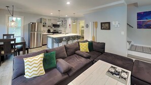 Eine moderne Küche mit Edelstahlgeräten, ein Essbereich mit einem dunklen Holztisch und Stühlen sowie ein Wohnzimmer mit einem großen, braunen Ecksofa.