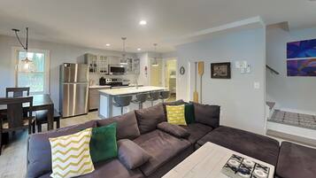 A modern kitchen with stainless steel appliances, a dining area with a dark wood table and chairs, and a living room with a large brown sectional sofa.