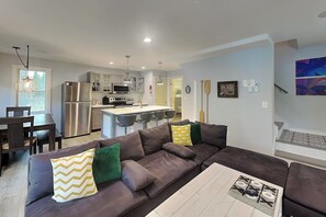 Eine moderne Küche mit Edelstahlgeräten, ein Essbereich mit einem dunklen Holztisch und Stühlen sowie ein Wohnzimmer mit einem großen, braunen Ecksofa.