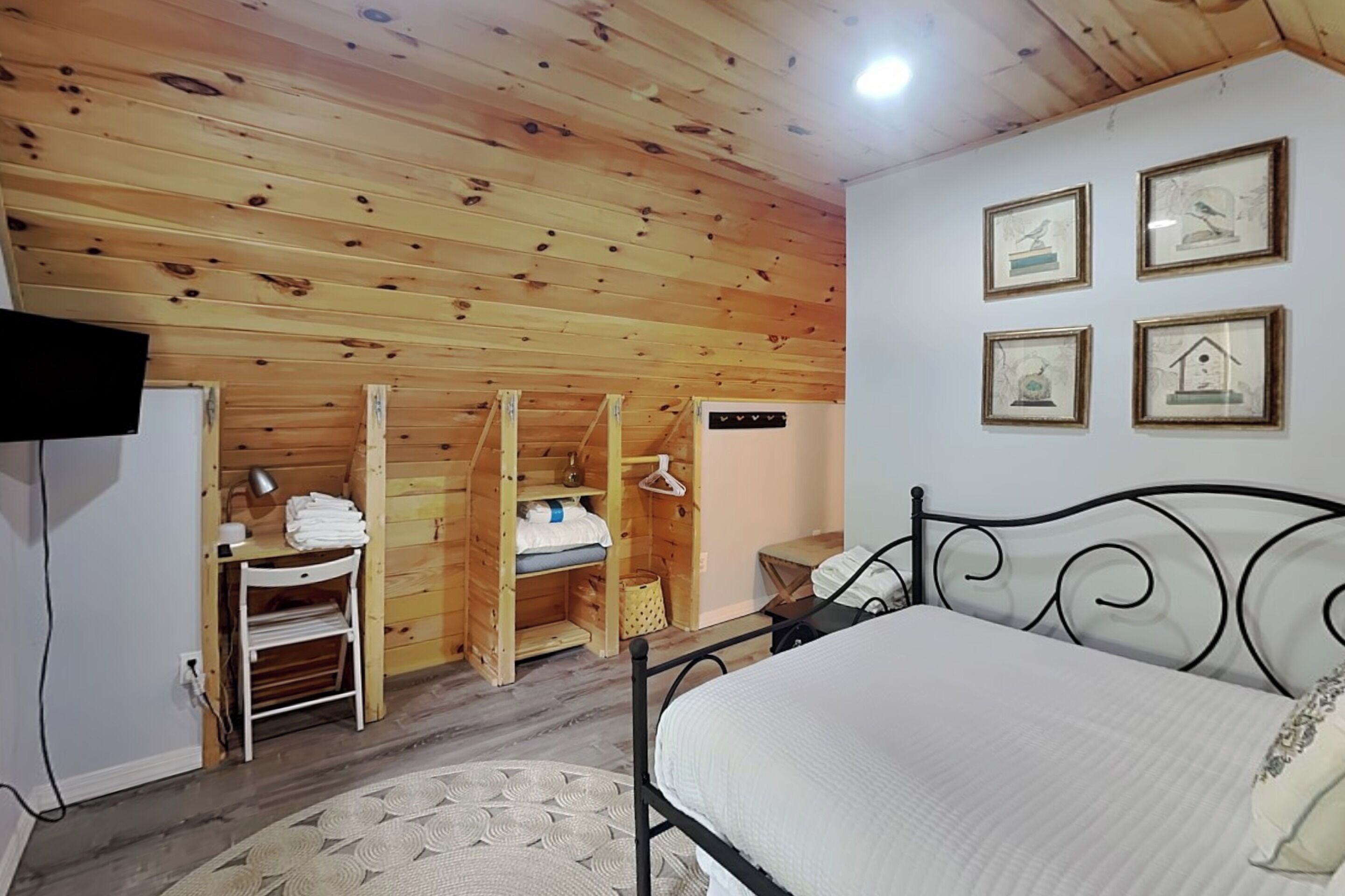 Une chambre avec un plafond en bois, un lit avec un matelas blanc, un téléviseur fixé au mur et une petite table avec une lampe.