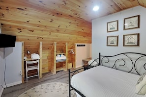 A bedroom with a wooden ceiling, a bed with a white mattress, a TV mounted on the wall, and a small table with a lamp.