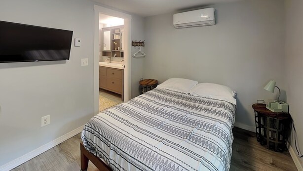 Ein Schlafzimmer mit einem Bett, einem an der Wand befestigten Fernseher, einem Nachttisch mit Lampe und einer Klimaanlage.