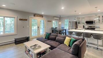 A modern living room with a sectional sofa, a coffee table, and a dining area with bar stools. There is a kitchen with a microwave and a refrigerator.
