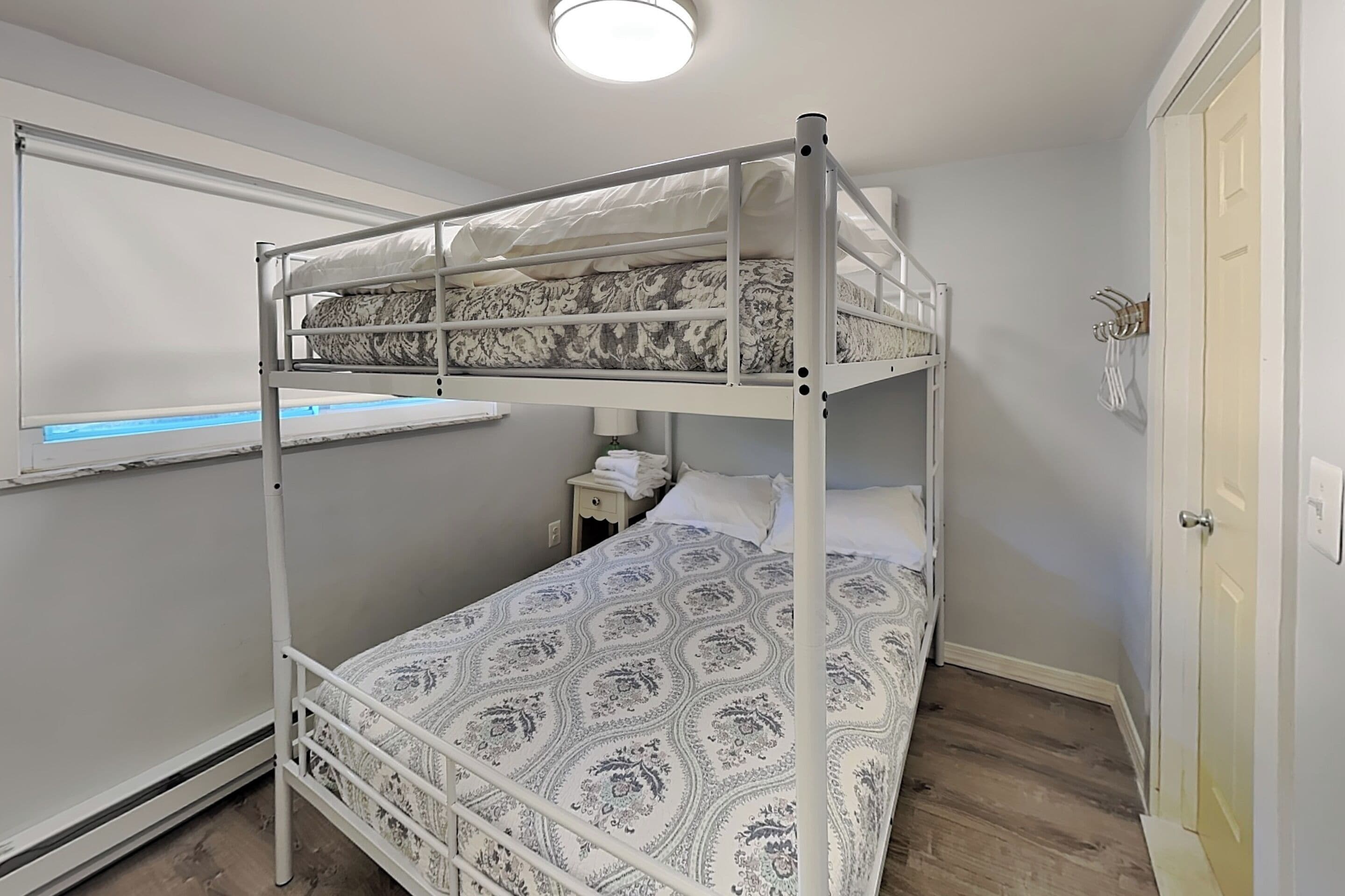 A bunk bed with a patterned bedspread, a bedside table with a lamp, and a window with a view of the outside.