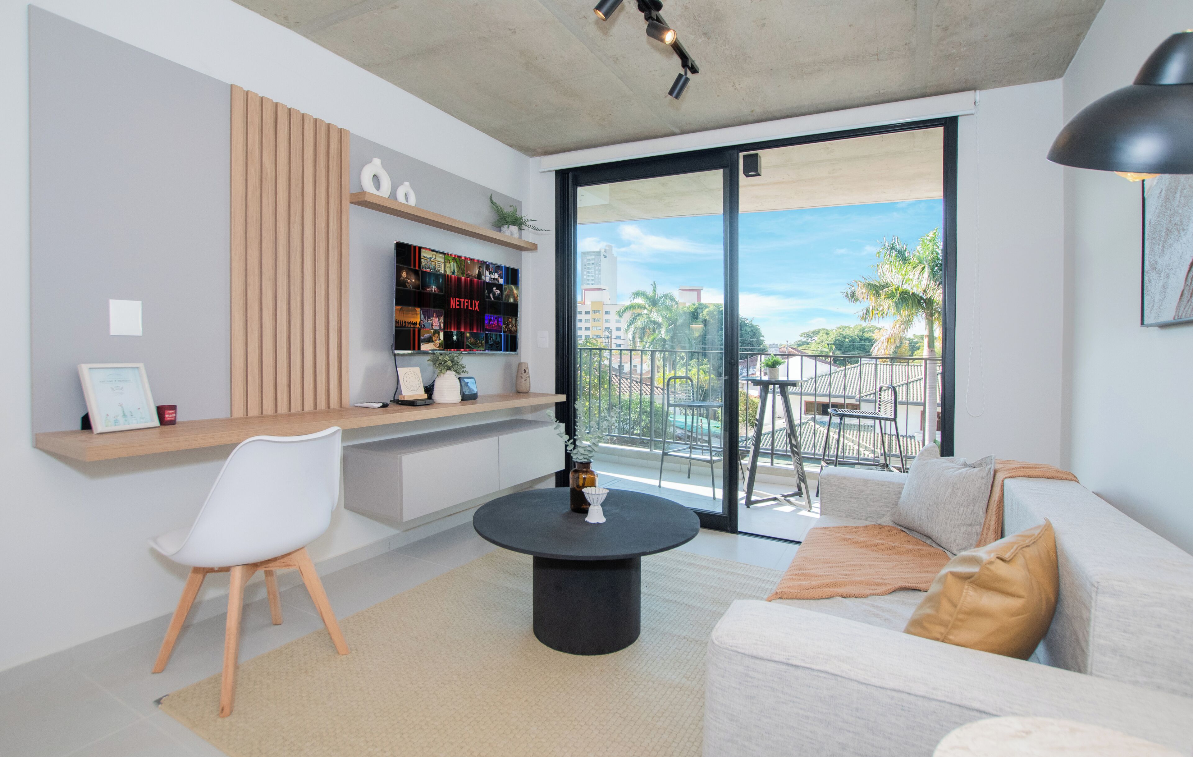 A modern living room with a sofa, a coffee table, a TV mounted on a wooden panel, and a balcony with a view of palm trees.