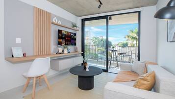 A modern living room with a sofa, a coffee table, a TV mounted on a wooden panel, and a balcony with a view of palm trees.