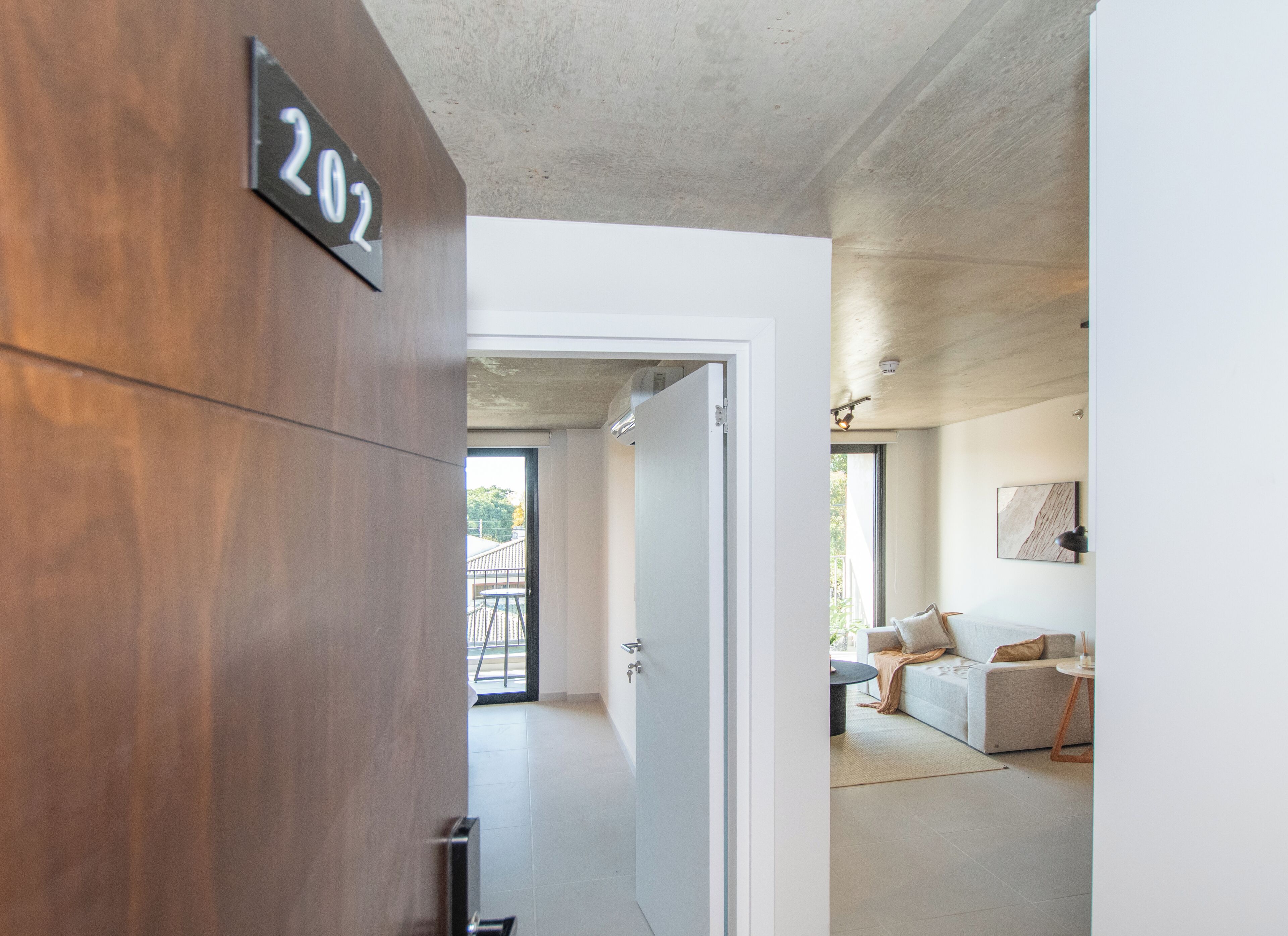 A modern interior with a wooden door marked '202', a white wall, and a glimpse of a living area with a sofa and a table.