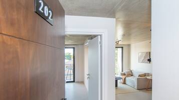 A modern interior with a wooden door marked '202', a white wall, and a glimpse of a living area with a sofa and a table.