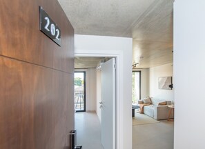 Un interior moderno con una puerta de madera marcada '202', una pared blanca y una vista de una sala de estar con un sofá y una mesa.