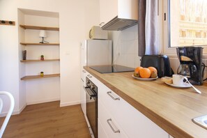 A kitchen with wooden cabinets, a built-in oven, a coffee maker, and a window with a view of bamboo. - La Maisonnette de Cholet - Studio avec jardin (Cholet)