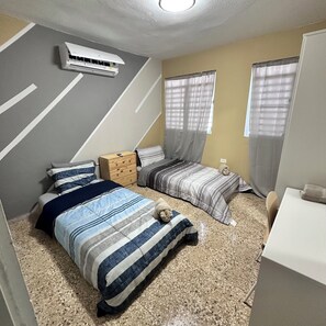 Een slaapkamer met twee bedden, een houten nachtkastje, een wandairconditioner en een raam met gordijnen.