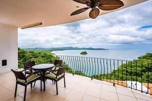 A balcony with a table and chairs, a ceiling fan, and a view of a body of water with greenery and distant islands. - Stylish Oceanview Condo with Quick Beach Access, Modern Design & Spacious Balcony (Santa Cruz)