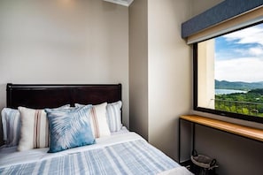A bedroom with a bed, pillows, a window with a view of a landscape, and a small bedside table. - Stylish Oceanview Condo with Quick Beach Access, Modern Design & Spacious Balcony (Santa Cruz)