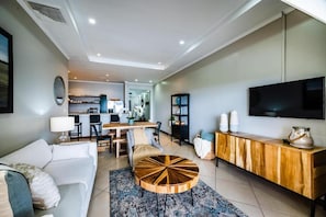 A modern living room with a sofa, a round coffee table, a TV, and a dining area with chairs and a table. - Stylish Oceanview Condo with Quick Beach Access, Modern Design & Spacious Balcony (Santa Cruz)