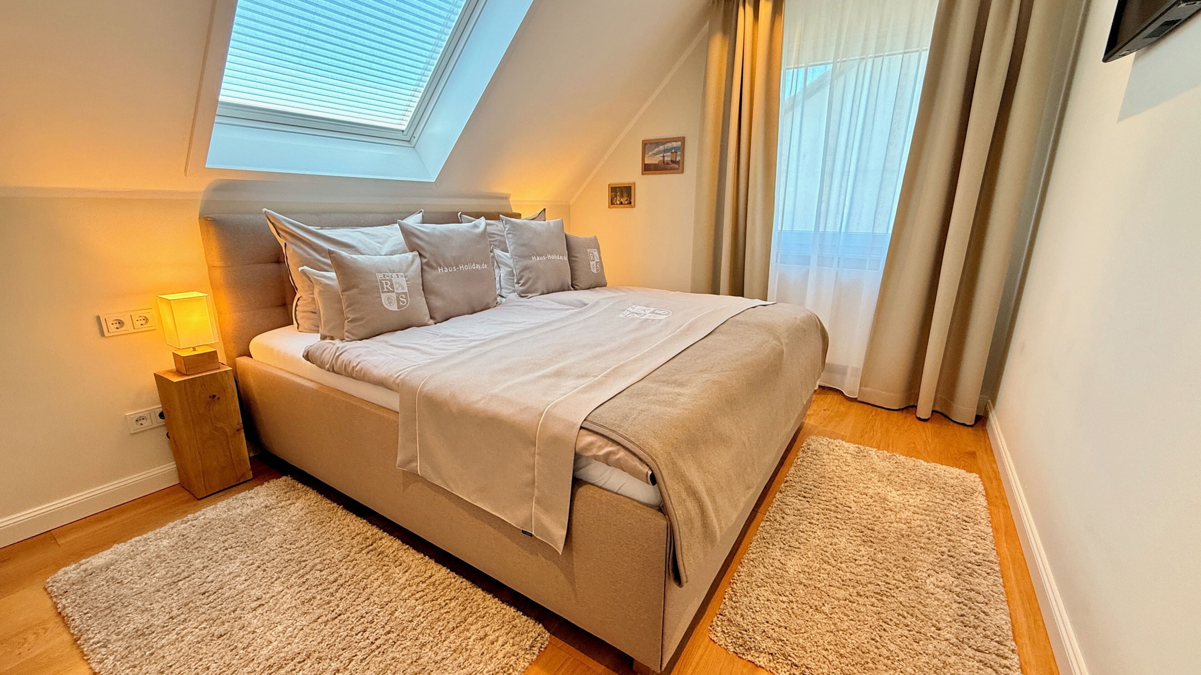 Une chambre à coucher comprenant un lit, une table de chevet, une lampe, une fenêtre avec des stores et une vue sur l’extérieur.