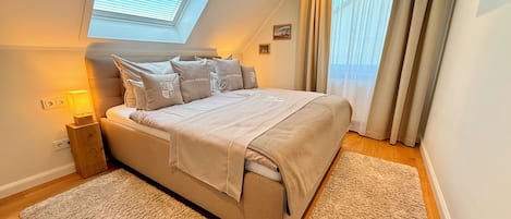 A bedroom with a bed, a nightstand, a lamp, a window with blinds, and a view of the outside.