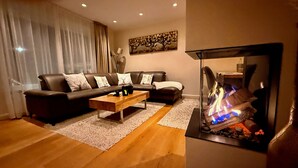 Ein Wohnzimmer mit einer Sofagarnitur, einem Couchtisch, einem Kamin mit Feuer und einem Bild an der Wand.