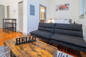 A modern living room with a dark sofa, a wooden coffee table with 'HELLO' letters, a small side table, and a painting on the wall. - Large Studio - Nearby Parking - Hospitals - 3F (Louisville)