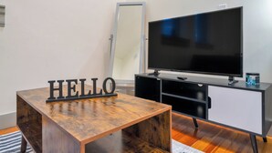Ein Zimmer mit einem Holztisch, einem schwarzen 'HELLO'-Schild, einem Flachbildfernseher und einem schwarzen Schrank.