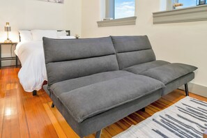 A gray sofa in a room with wooden flooring, a bed with white bedding, a nightstand with a lamp, and a window with a view. - Large Studio - Nearby Parking - Hospitals - 3F (Louisville)