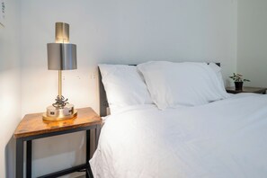 Una cama bien hecha con sábanas blancas, una mesita de noche con lámpara y una pequeña planta en maceta.