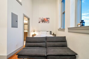 A hotel room with a bed, a bedside table, a lamp, a window with a view, and a door leading to a bathroom. - Large Studio - Nearby Parking - Hospitals - 3F (Louisville)