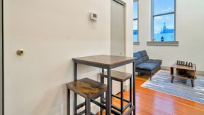 Ein Zimmer mit einem Holztisch, einem Stuhl, einem kleinen Tisch mit Lampe und Blick auf ein Gebäude durch ein Fenster.
