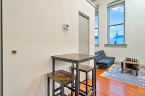 A room with a wooden table, a chair, a small table with a lamp, and a view of a building through a window. - Large Studio - Nearby Parking - Hospitals - 3F (Louisville)