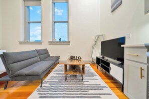 A modern living room with a grey sofa, a wooden coffee table with a 'HELLO' sign, a flat-screen TV, and a white cabinet. - Large Studio - Nearby Parking - Hospitals - 3F (Louisville)