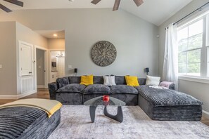 A living room with a sectional sofa, a glass coffee table, and a decorative wall piece. - Convenient New Modern House - Tidy and Equipped (Spartanburg)