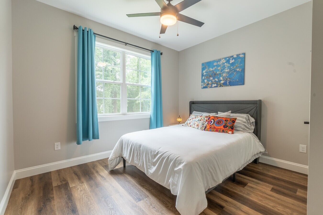 A bedroom with a bed, a ceiling fan, a window with curtains, and a painting on the wall.