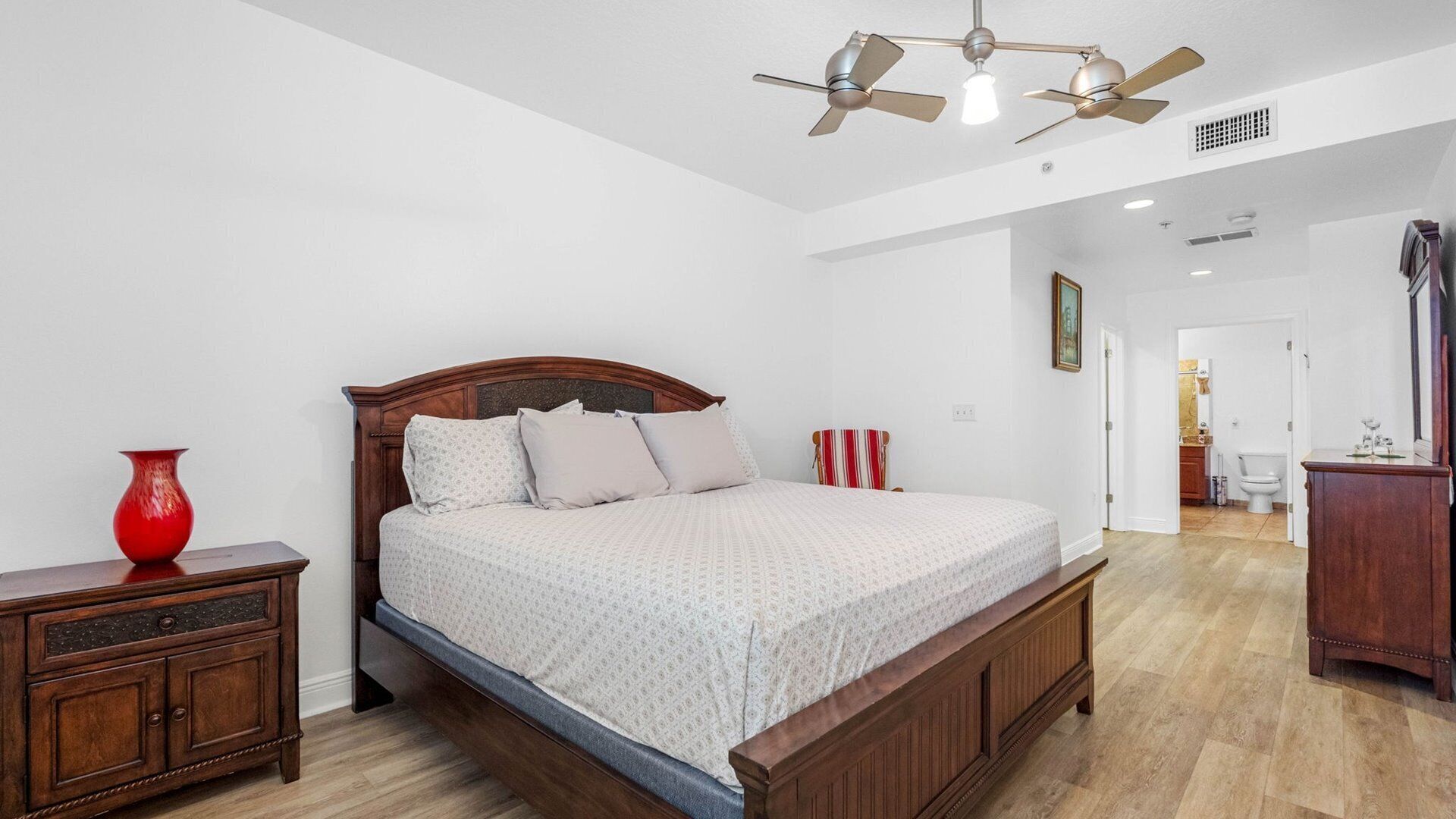 Une chambre à coucher avec un lit en bois, une table de chevet, un vase rouge et un ventilateur de plafond.