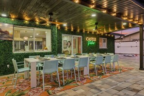 Ein Essbereich im Freien mit einem langen Tisch und Stühlen, umgeben von einer grünen Wand mit einem Neonlicht, unter einer Holzbalkendecke mit Lichterketten.