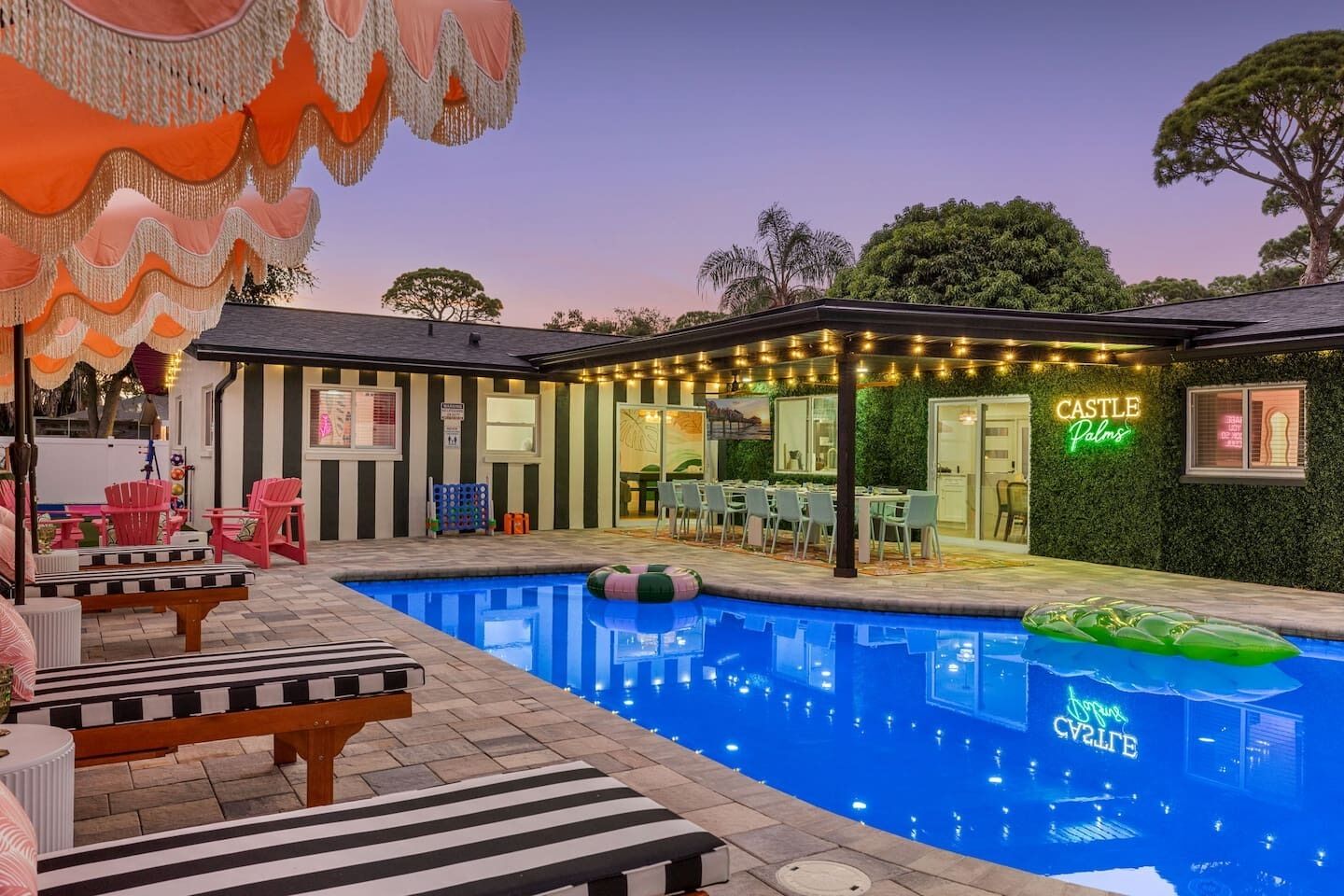 Un espace piscine avec des chaises longues, un coin repas et un panneau lumineux néon indiquant « CASTLE Palms ».