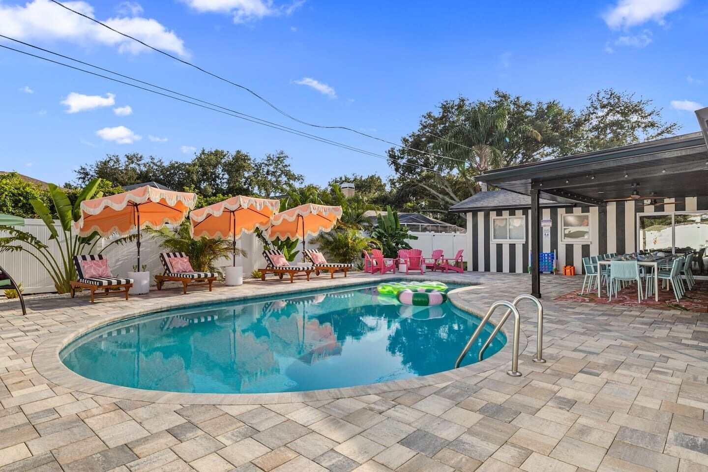 A swimming pool with lounge chairs and umbrellas, a covered outdoor seating area, and a view of trees and a building in the background.
