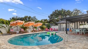 A swimming pool with lounge chairs and umbrellas, a covered outdoor seating area, and a view of trees and a building in the background.