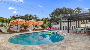 A swimming pool with lounge chairs and umbrellas, a covered outdoor seating area, and a view of trees and a building in the background.
