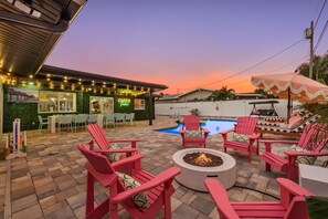 An outdoor patio area with a pool, red Adirondack chairs, a fire pit, and a bar with string lights.