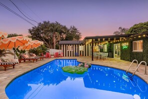 Ein Schwimmbecken mit Liegestühlen und Sonnenschirmen, ein kleines Gebäude mit einer Neonreklame und eine Terrasse mit Lichterketten.