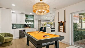 A modern indoor pool table with a pool cue, surrounded by a kitchen area with white cabinets and a green backsplash.